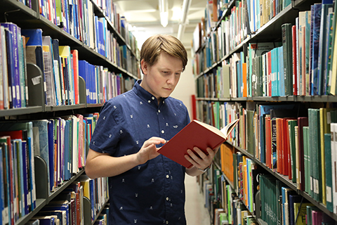 This is a photo of an English major at the University of Miami in the library. The student is in one of the aisles with an open book in their hand. The student appears to be reading the book.